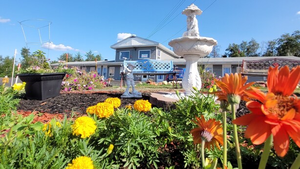 A garden with a white fountain, yellow and orange flowers, and a building with a blue roof in the background. - Norvic Motel (Sudbury)