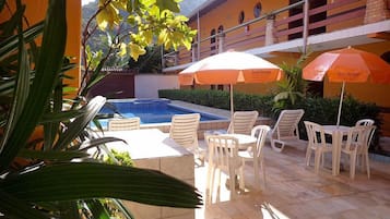 Zona de piscina al aire libre con sillas de plástico blancas y sombrillas naranjas, rodeada de vegetación y un edificio de paredes amarillas.