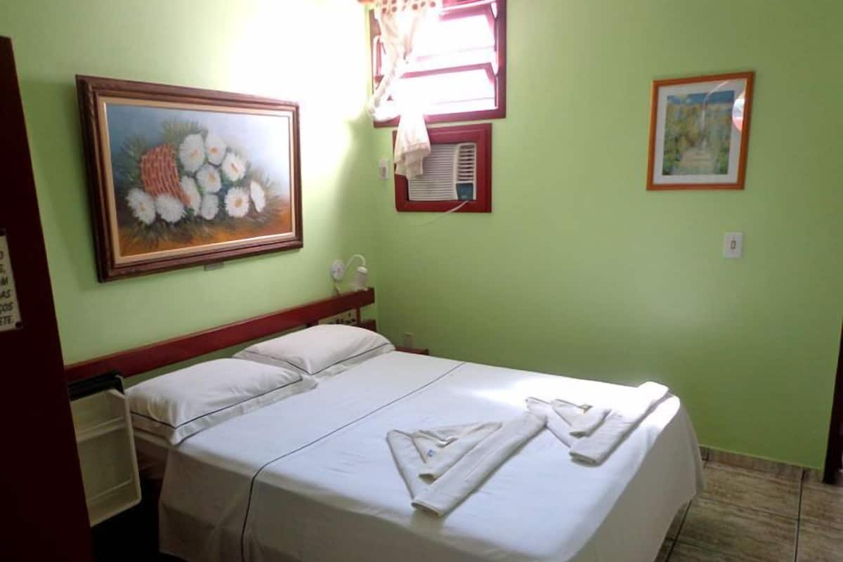 A neatly made bed with white linens and towels, a painting on the wall, and a window with blinds.