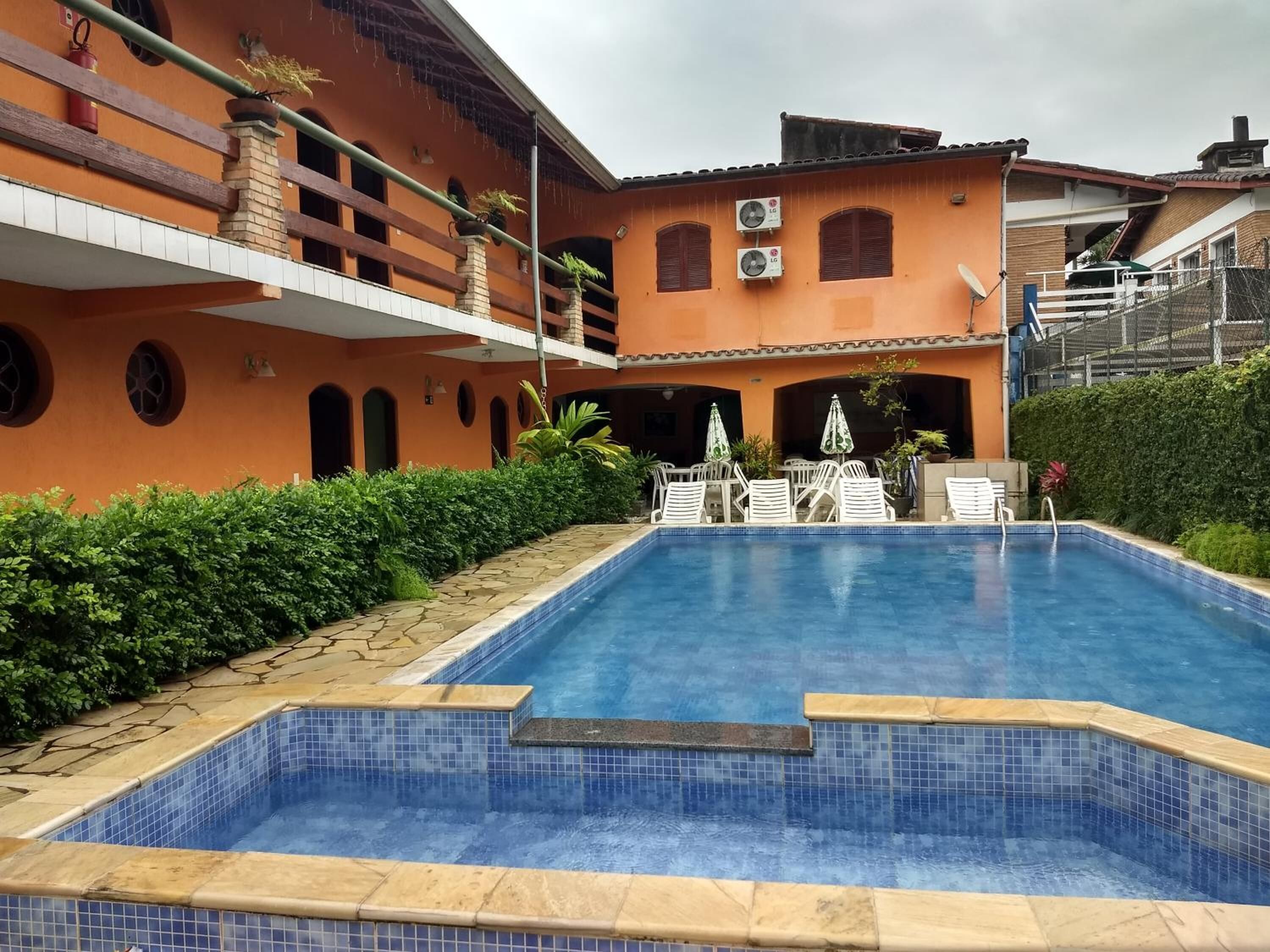 Una piscina con borde de azulejos, rodeada de un seto y un edificio de fachada naranja y múltiples ventanas.
