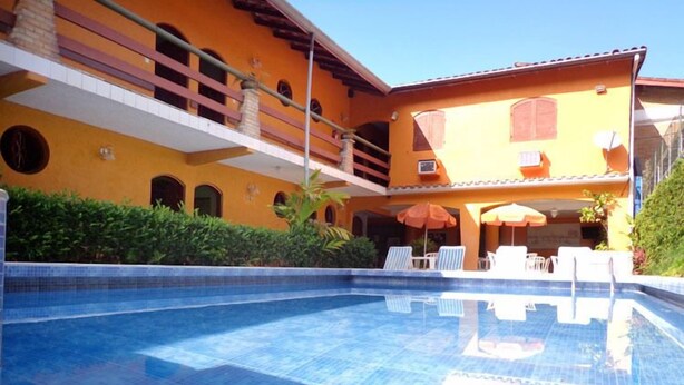 A swimming pool with lounge chairs and umbrellas, surrounded by a building with a yellow facade and balconies. - Hotel Pousada Miramar (Ubatuba)