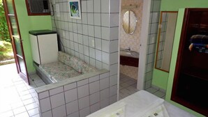 A small, tiled bathroom with a shower, a sink, and a toilet. There is a bench with a floral pattern and a small window with a picture. - Hotel Pousada Miramar (Ubatuba)