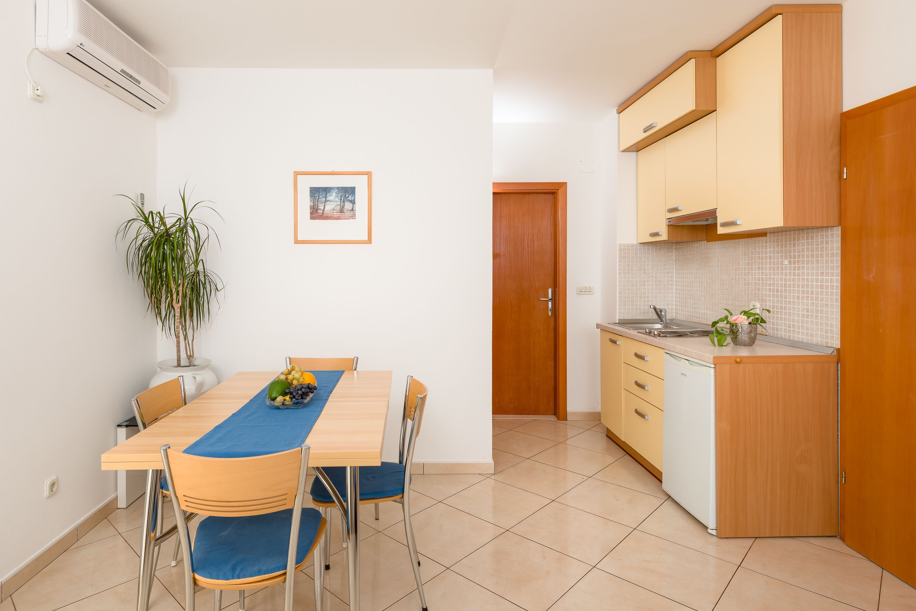 A compact kitchen with a dining area, featuring a wooden table, chairs, a potted plant, and a tiled backsplash.