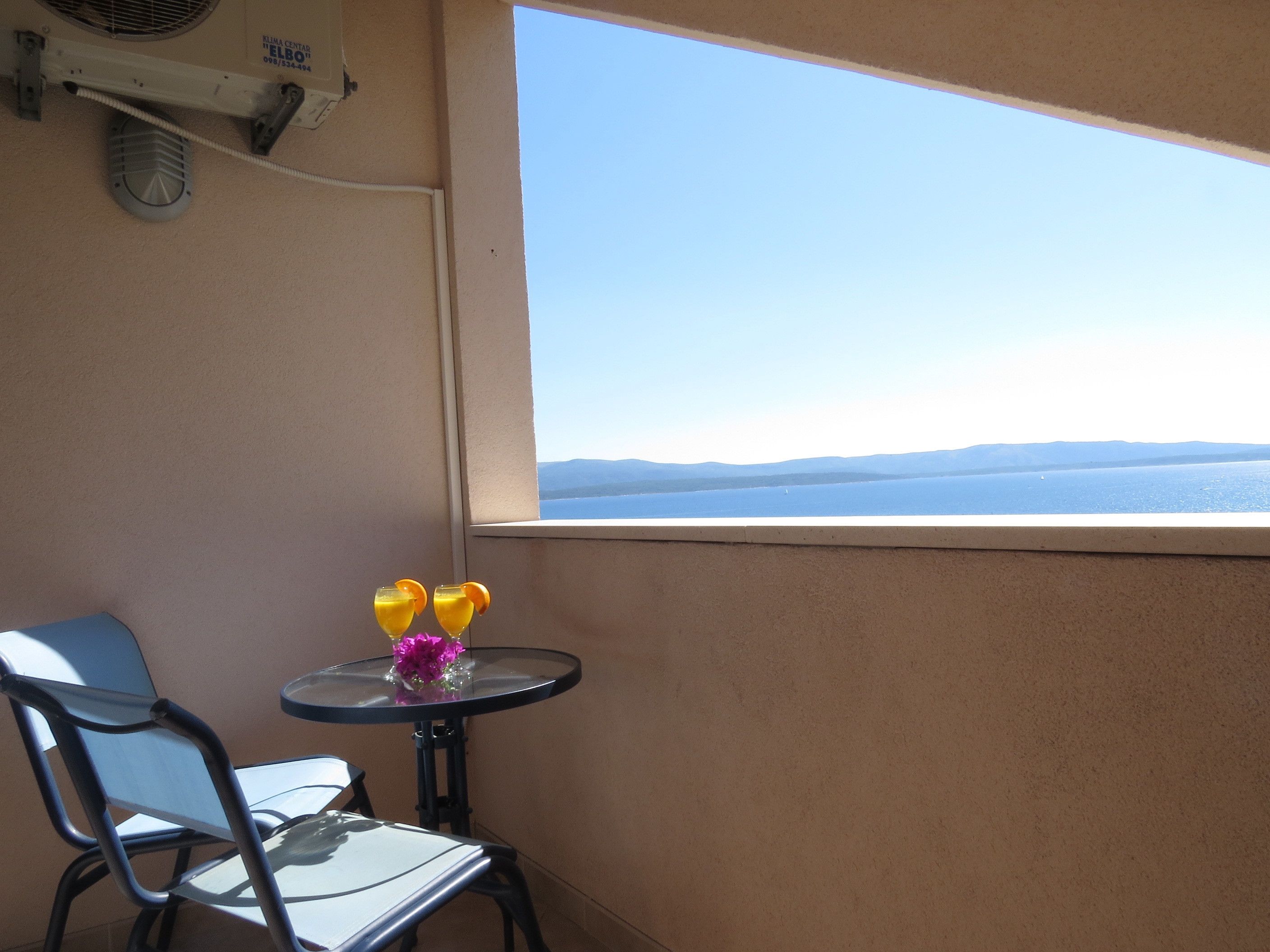 A balcony with a table and chairs, a window with a view of the sea and mountains, and a wall-mounted air conditioning unit.