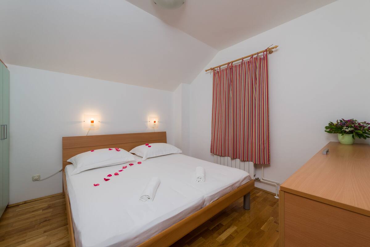 Una cama impecablemente arreglada con pétalos de rosa y toallas, un cabecero de madera, una mesita de noche y una ventana con cortina a rayas.