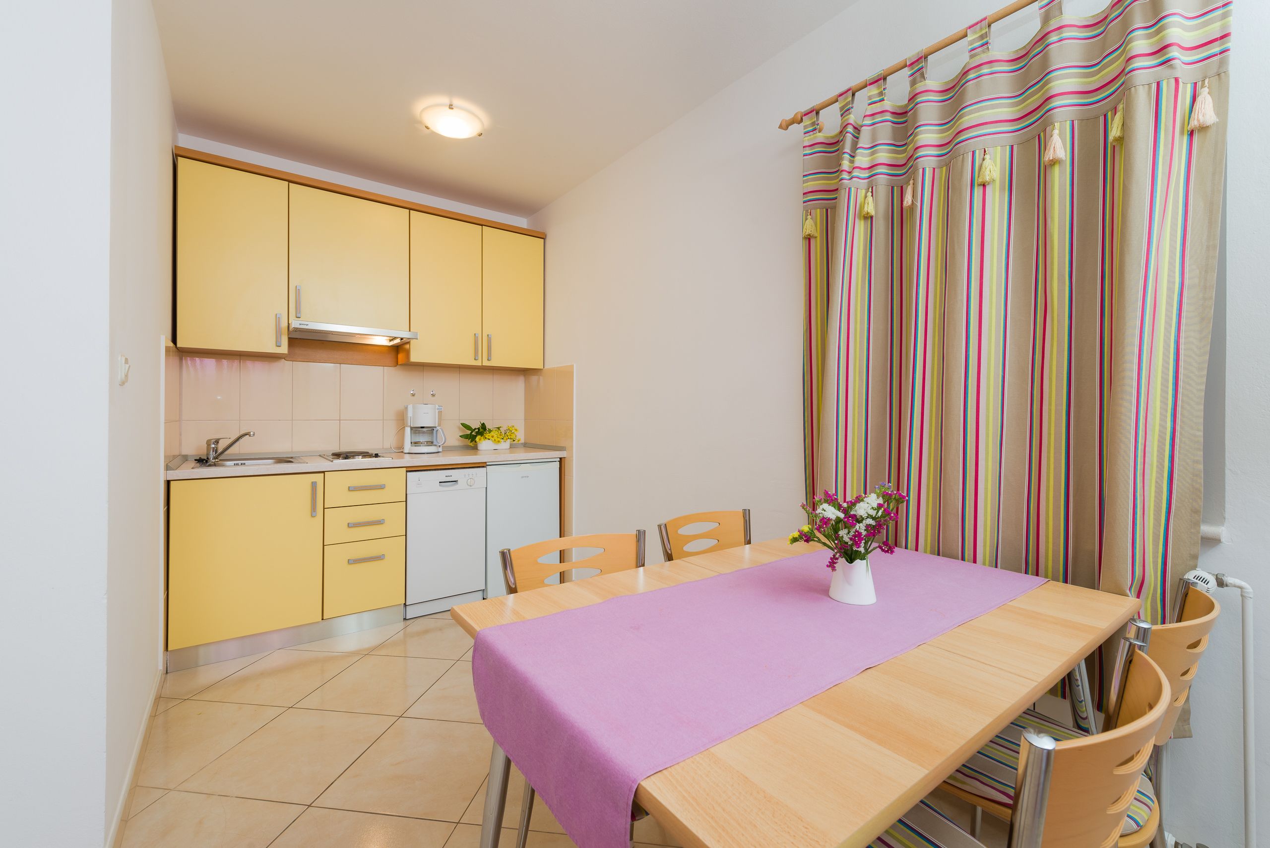 A compact kitchen with yellow cabinets, a wooden dining table with a purple tablecloth, and a striped curtain.