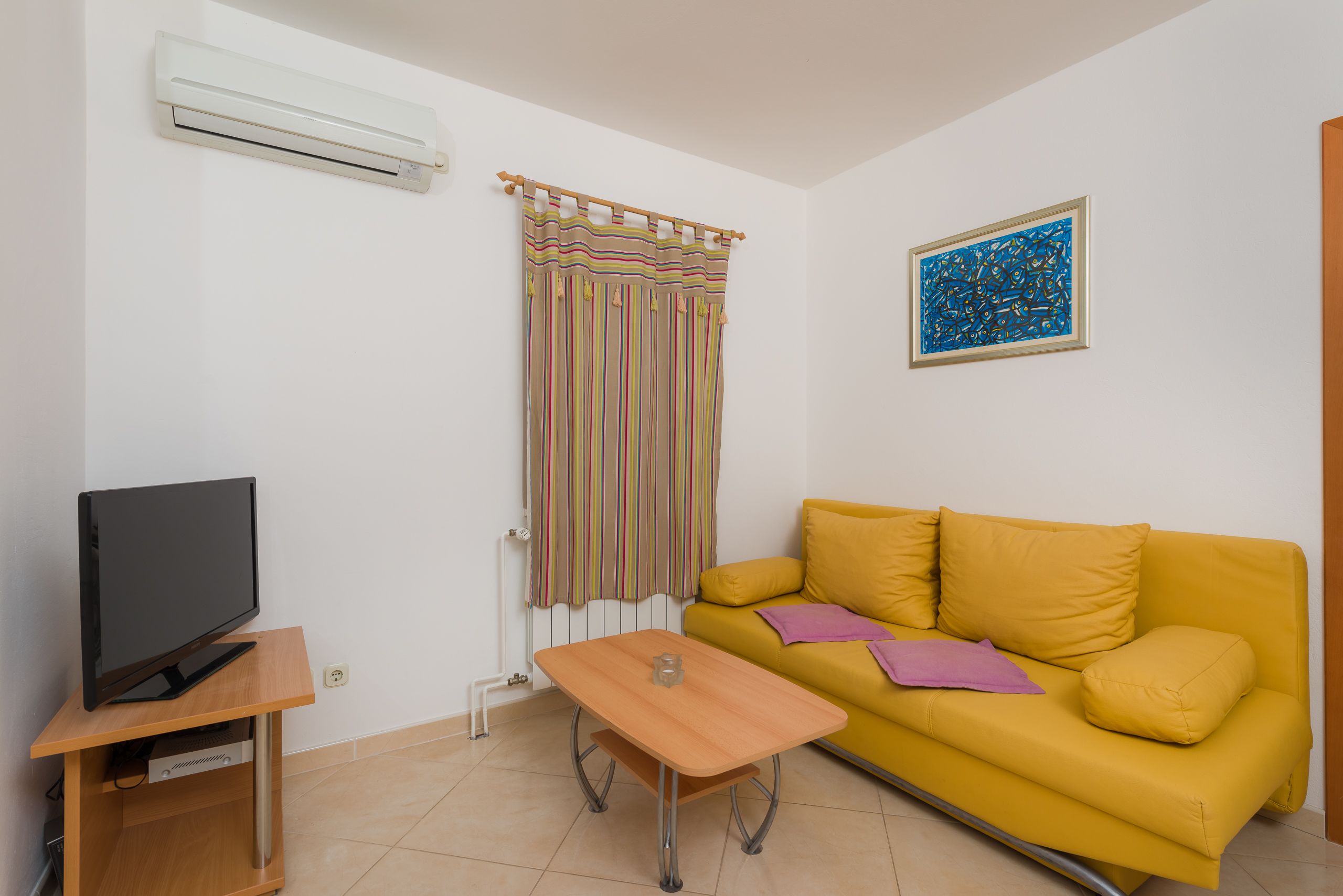 A living room with a yellow sofa, a TV on a wooden stand, a small table, a striped curtain, and a blue abstract painting on the wall.