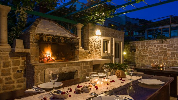 Un espace extérieur aménagé pour les repas, avec une cheminée en pierre, une table dressée pour deux personnes, agrémentée de verres à vin et de bougies, et une pergola au-dessus.