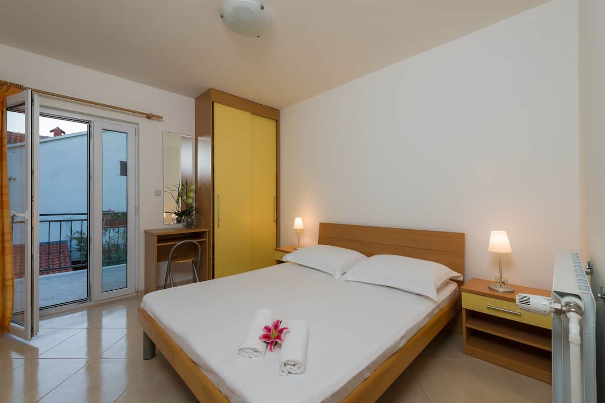Habitación de hotel con cama, mesitas de noche, escritorio, silla, puerta corrediza de vidrio al balcón y baño con toallero.