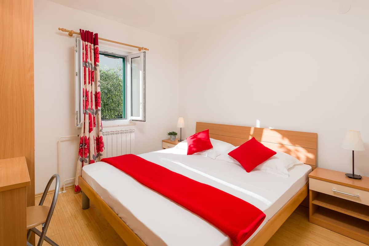 Una cama bien hecha con ropa de cama roja y blanca, un cabecero de madera y una mesita de noche con una lámpara.