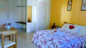 A hotel room with a bed, a table with a vase of flowers, a bedside table with a bottle and glasses, and a door leading to another room. - Pousada Porto do Sol (Cabo Frio)