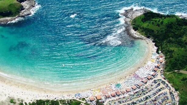 An aerial view of a beach with a curved shoreline, a large crowd of people, and a resort area with rows of beach umbrellas and tents. - Pousada Porto do Sol (Cabo Frio)