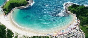 Vista aérea de una playa con una costa curvada, una gran multitud de personas y una zona de resort con filas de sombrillas y tiendas de playa.