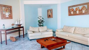 A living room with two beige sofas, a wooden coffee table, a small table with a ship model, and a potted plant. - Pousada Porto do Sol (Cabo Frio)