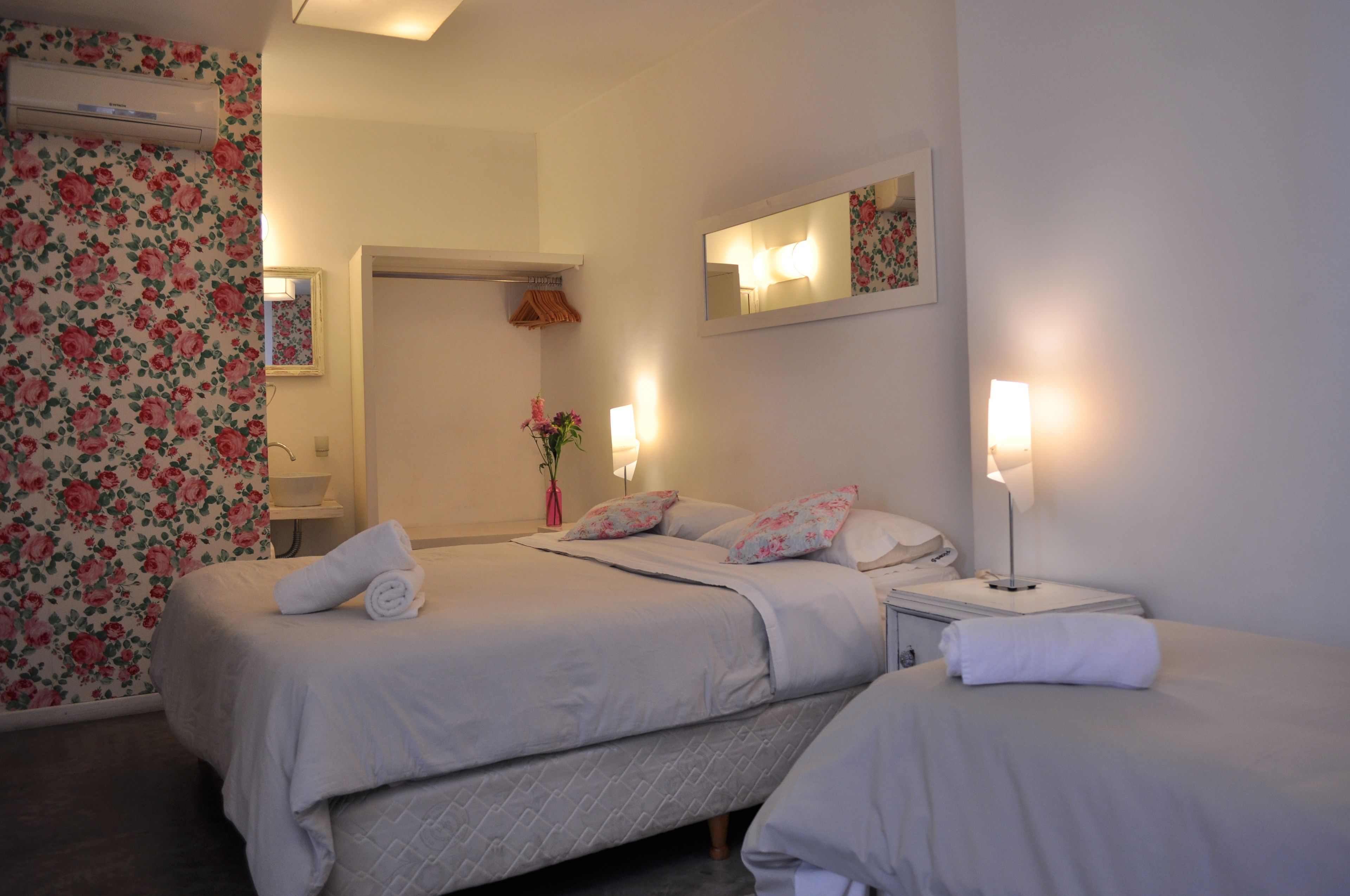 A hotel room with two beds, a mirror, a bedside table with a lamp, and a floral wallpaper on one wall.
