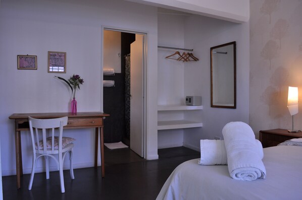 A hotel room with a bed, a desk with a chair, a wardrobe, a mirror, and a small bathroom area.