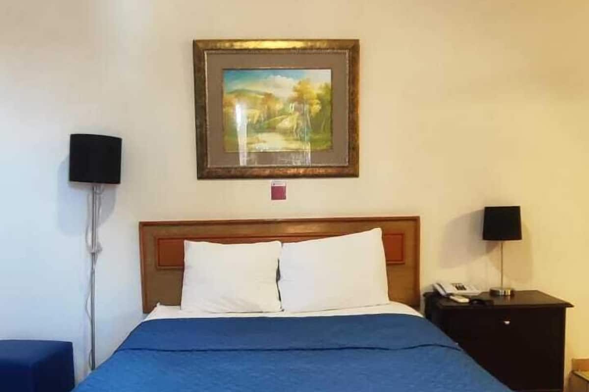 A hotel room with a large bed, a wooden headboard, two bedside tables with lamps, a framed picture on the wall, and a tiled floor.