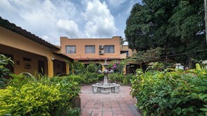 A courtyard with a central fountain, surrounded by lush greenery and a building with a tiled roof. - Hotel Mediterráneo Plaza (San Salvador)