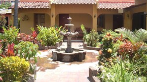 A courtyard with a central fountain, surrounded by lush greenery and a building with a tiled roof. - Hotel Mediterráneo Plaza (San Salvador)