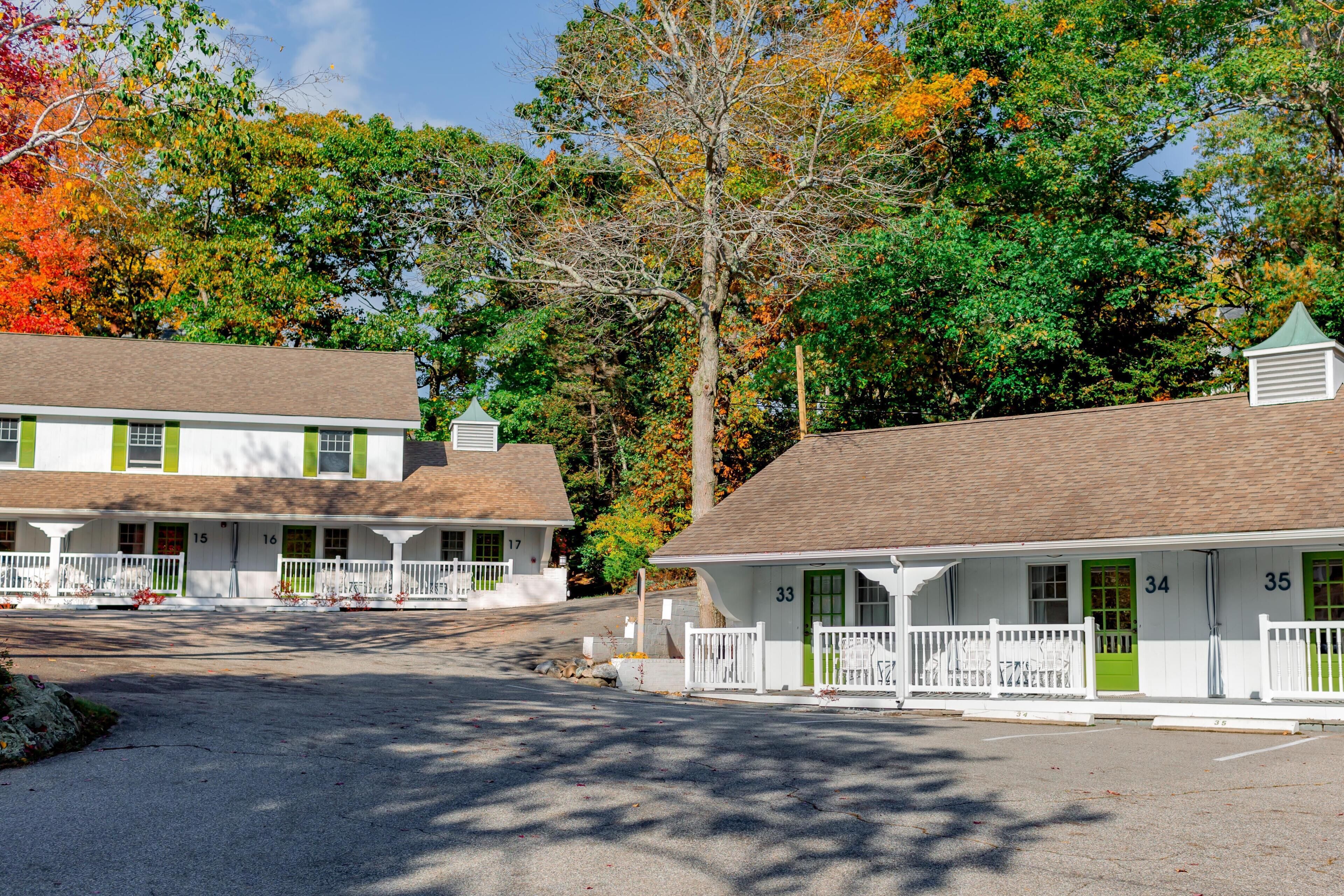 A row of white buildings with green doors and windows, numbered 15 to 35, set against a backdrop of autumn-colored trees.