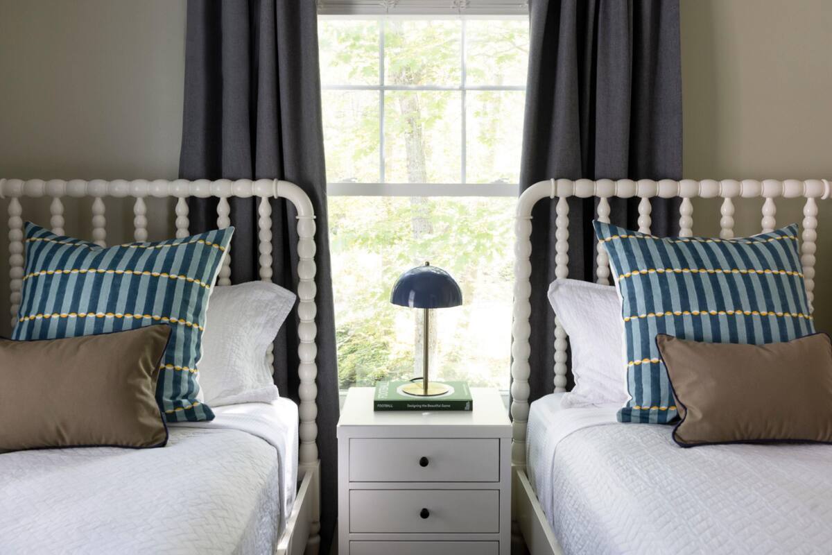 A bedroom with two beds, each with a white headboard, a bedside table with a lamp, and a window with curtains.