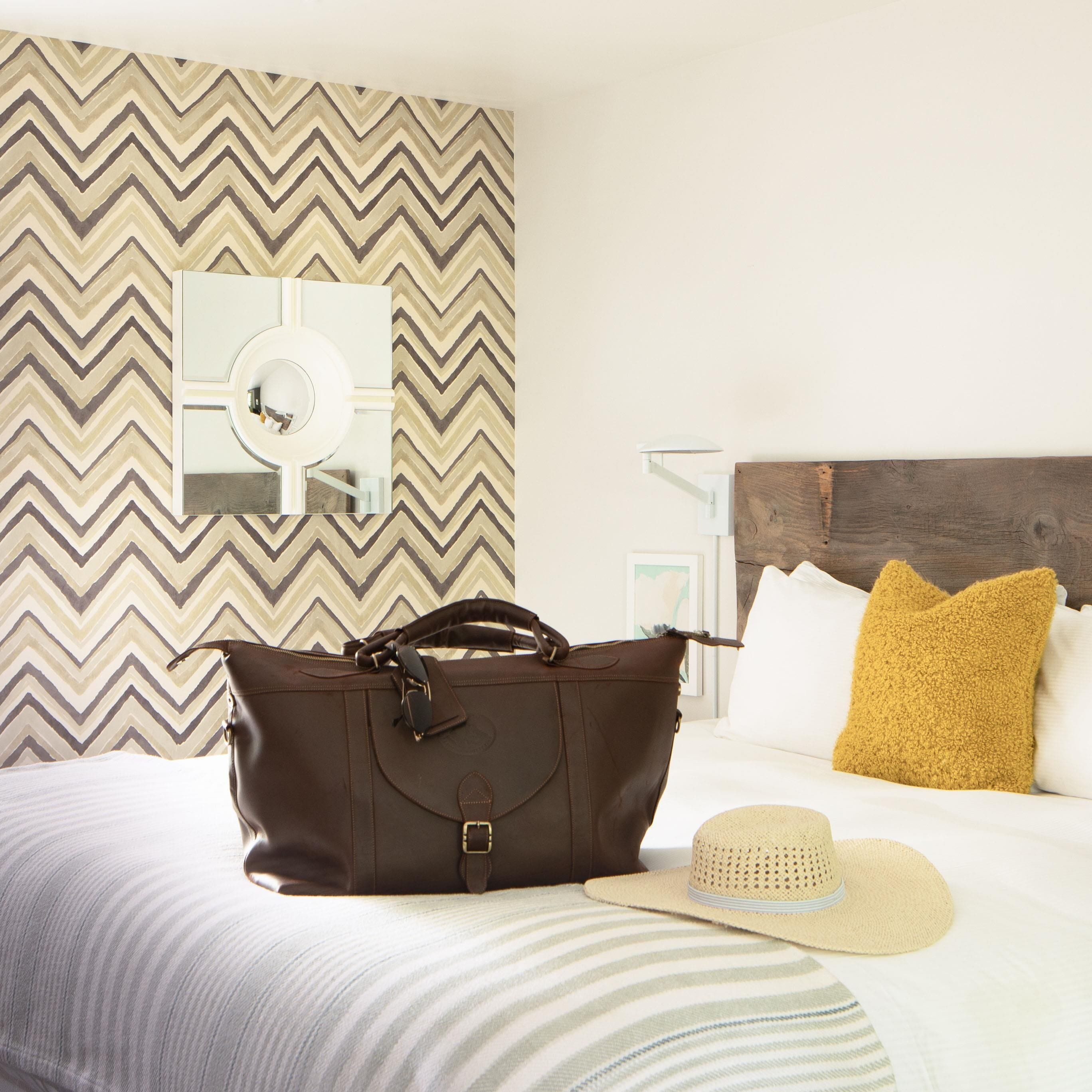 Habitación de hotel con una cama, un bolso de cuero marrón, un sombrero de paja y una pared con un diseño en zigzag.