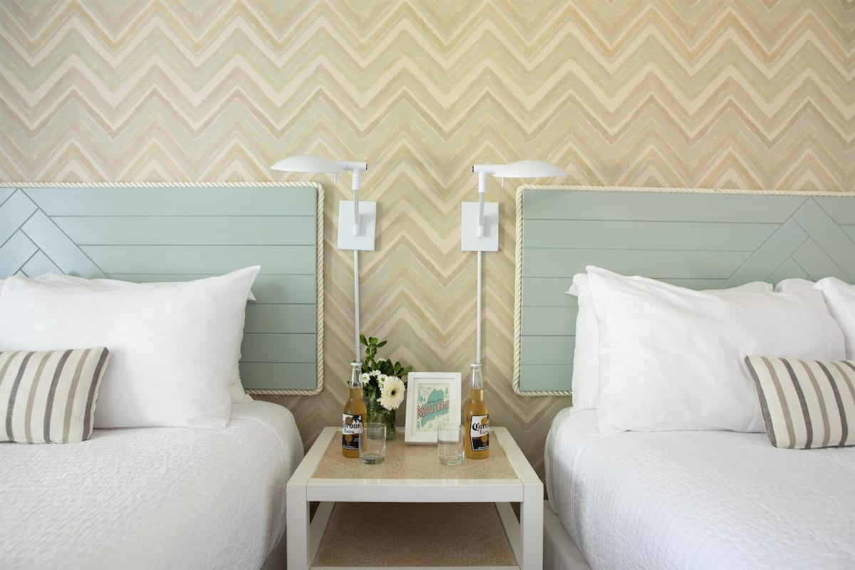 A hotel room with two beds, a small table with drinks and a framed picture, and a chevron-patterned wall.