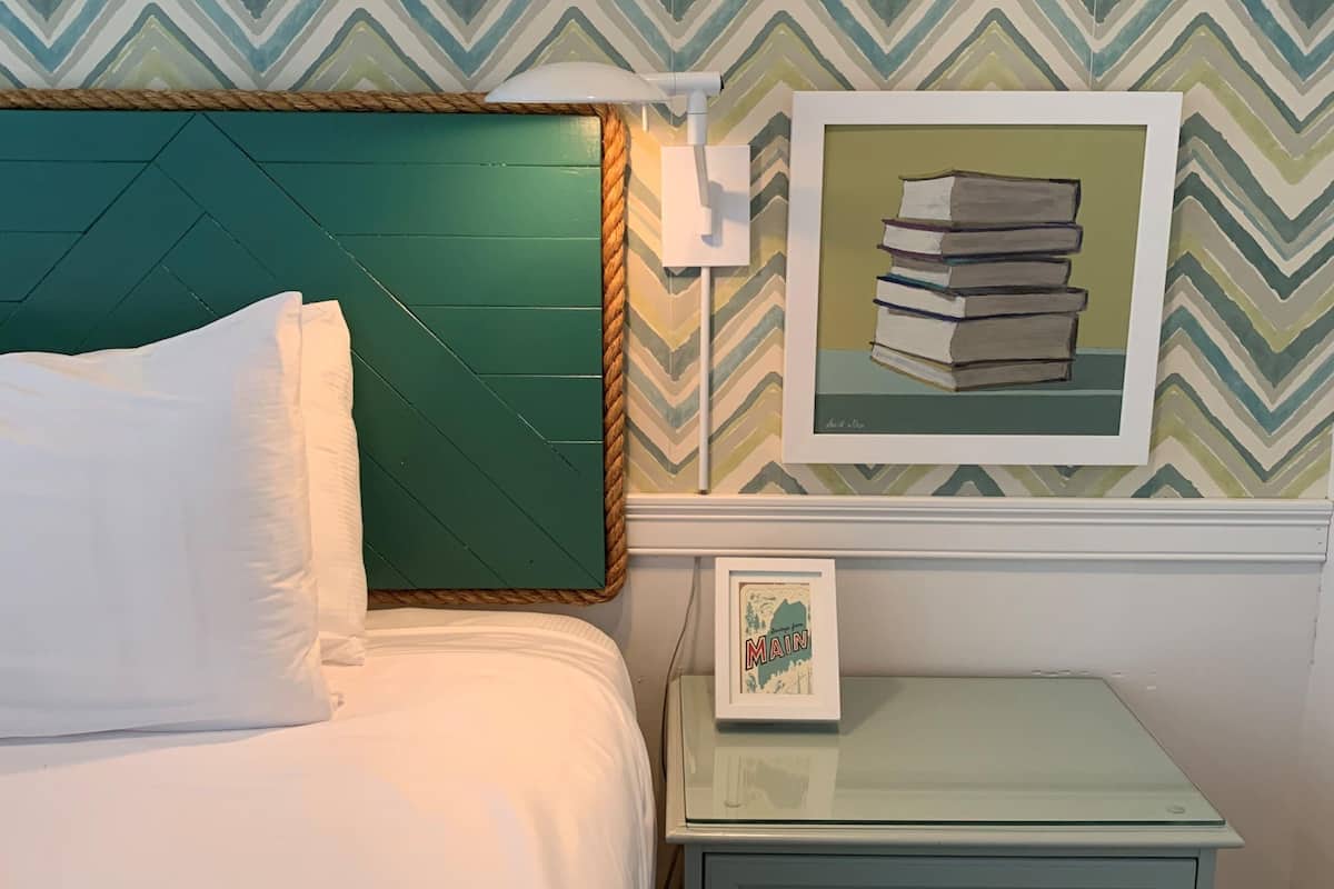 A bed with white linens, a green headboard, a framed picture of books, and a small bedside table with a drawer.