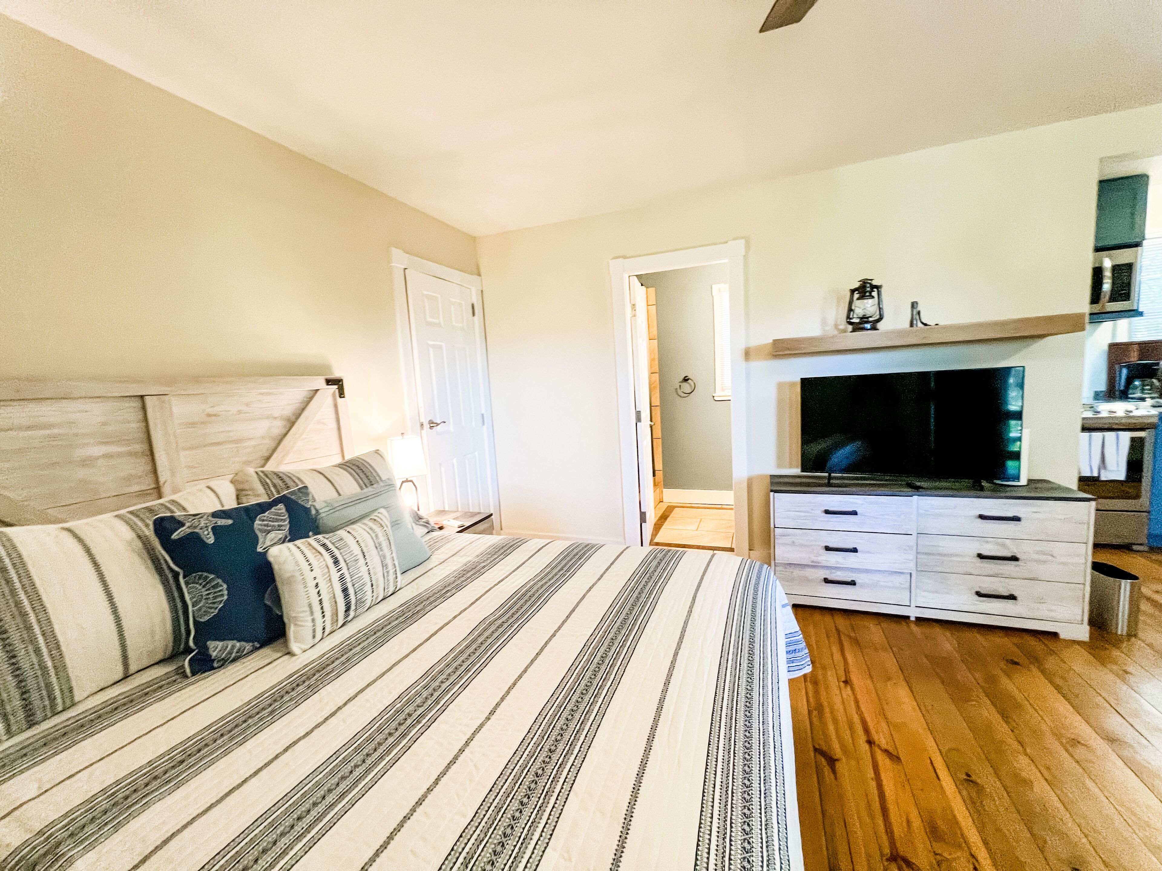 Un dormitorio con cama, cómoda, televisor y una zona de cocina al fondo.