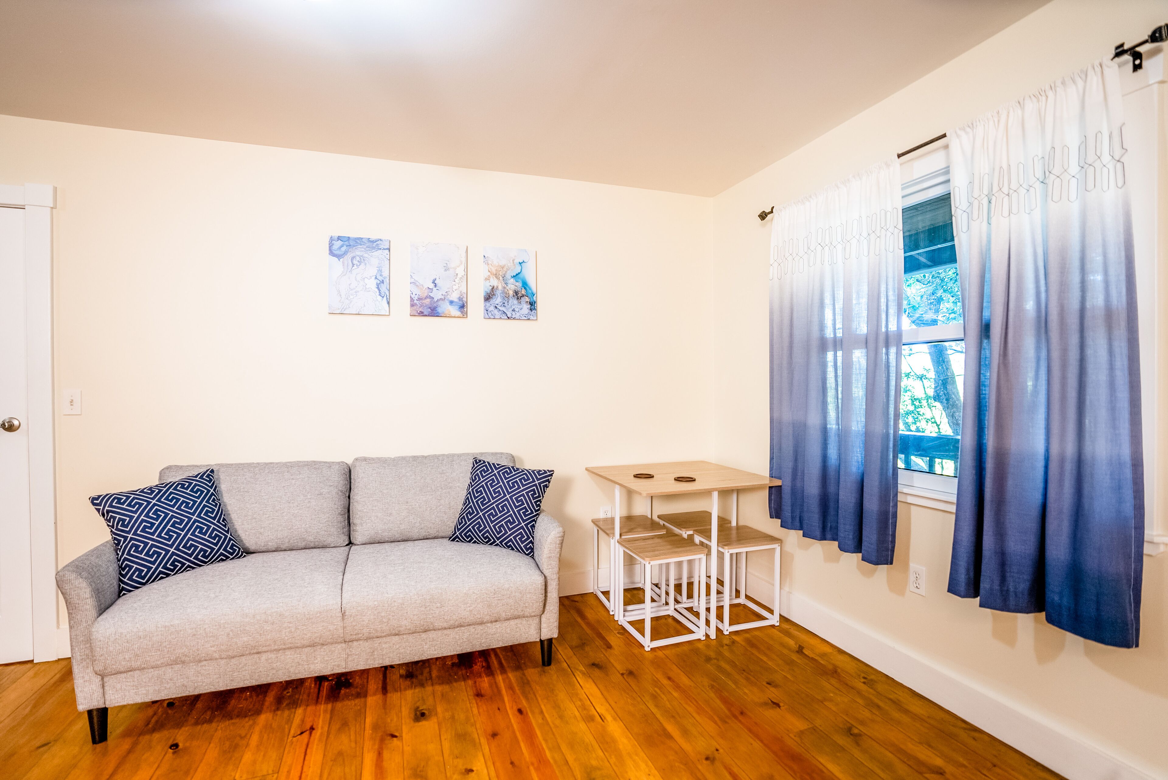 Ein Wohnzimmer mit grauem Sofa, zwei gemusterten Kissen, einem kleinen weißen Tisch und einem Fenster mit blauen Vorhängen.