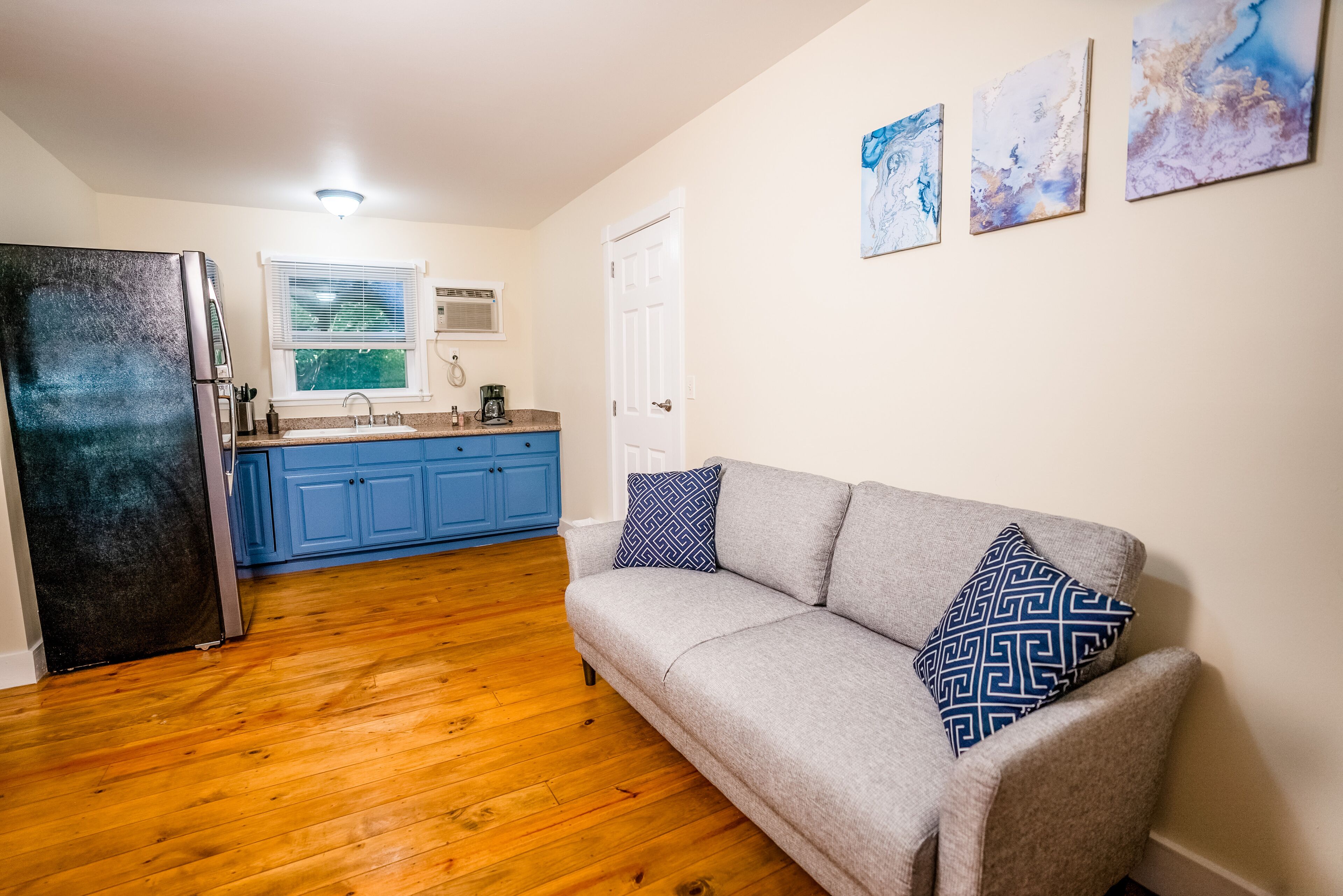 A living room with a grey sofa and patterned pillows, a wooden floor, a kitchen with blue cabinets, and a white door.