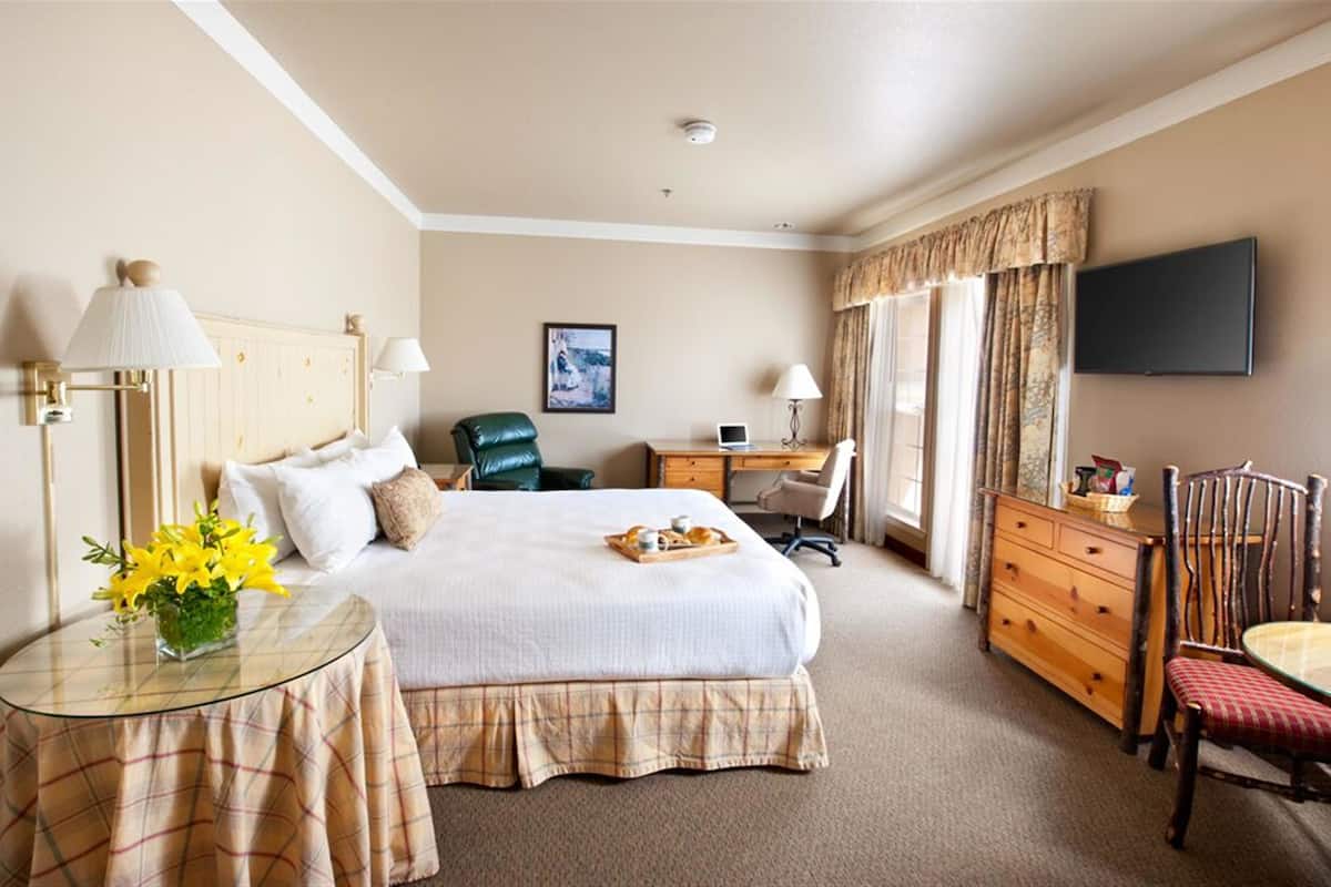 A hotel room with a bed, a desk, a chair, a dresser, a television, and a small table with flowers.