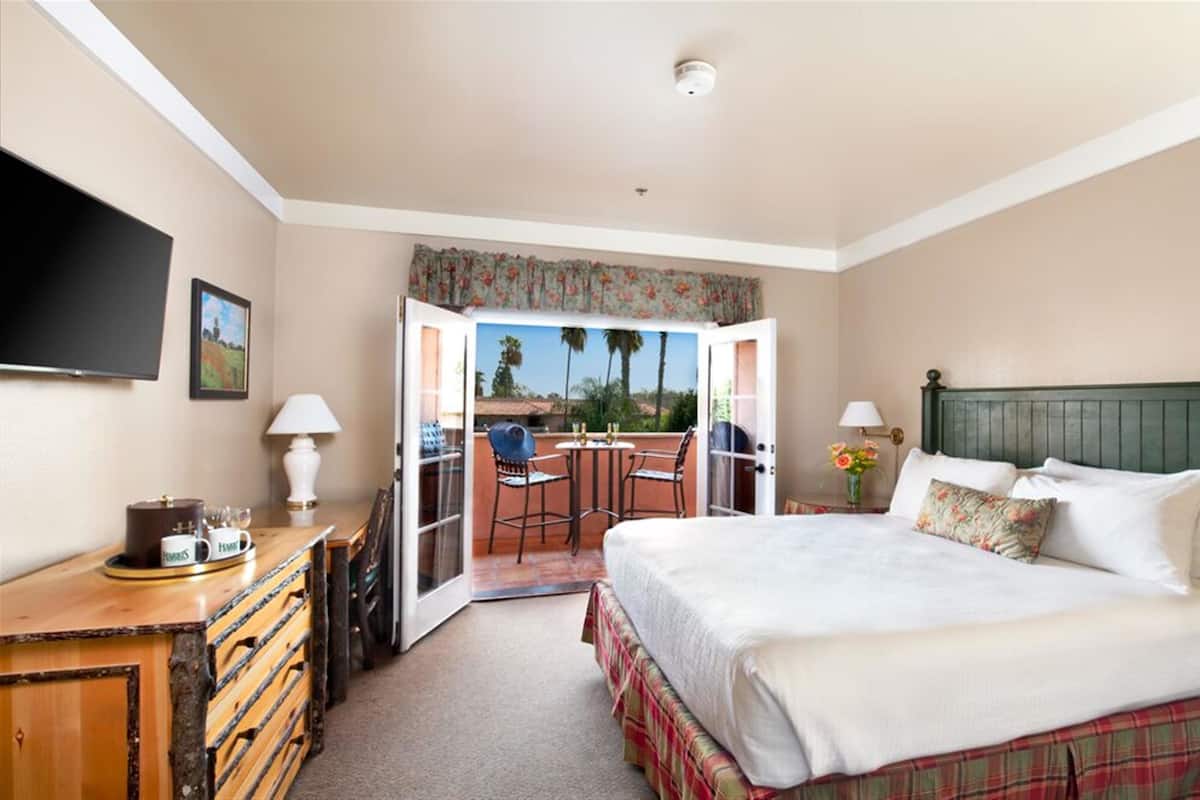 A hotel room with a bed, a desk with a lamp, a television, and a view of an outdoor patio with chairs and a table.