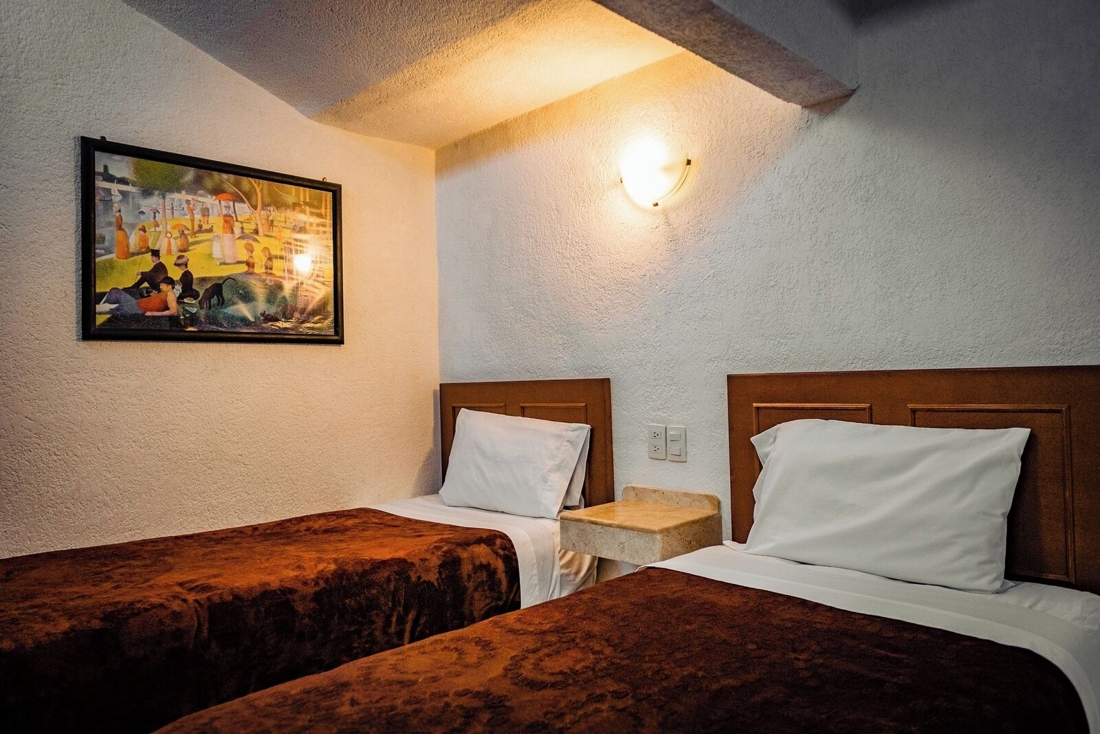 A hotel room with two single beds, a small bedside table, a wall-mounted light, and a painting on the wall.