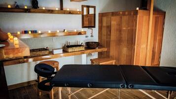 A sauna room with wooden benches, a bench with a massage table, shelves with candles, and a mirror.
