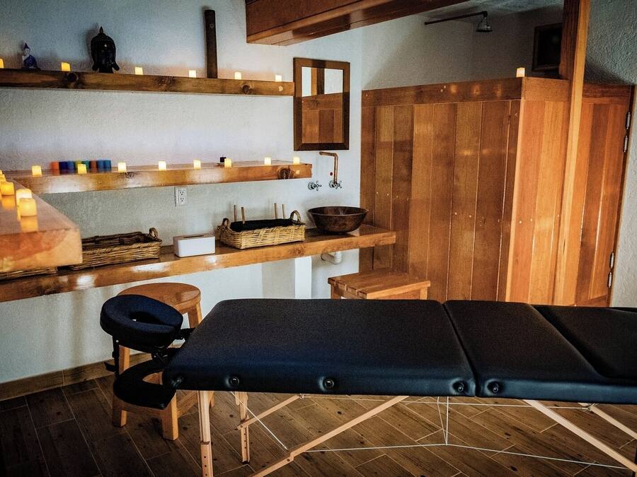 A sauna room with wooden benches, a bench with a massage table, shelves with candles, and a mirror.
