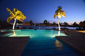 A swimming pool at dusk with palm trees and thatched umbrellas, surrounded by a calm water body and distant buildings. - Spanish Water Beach (Jan Thiel)