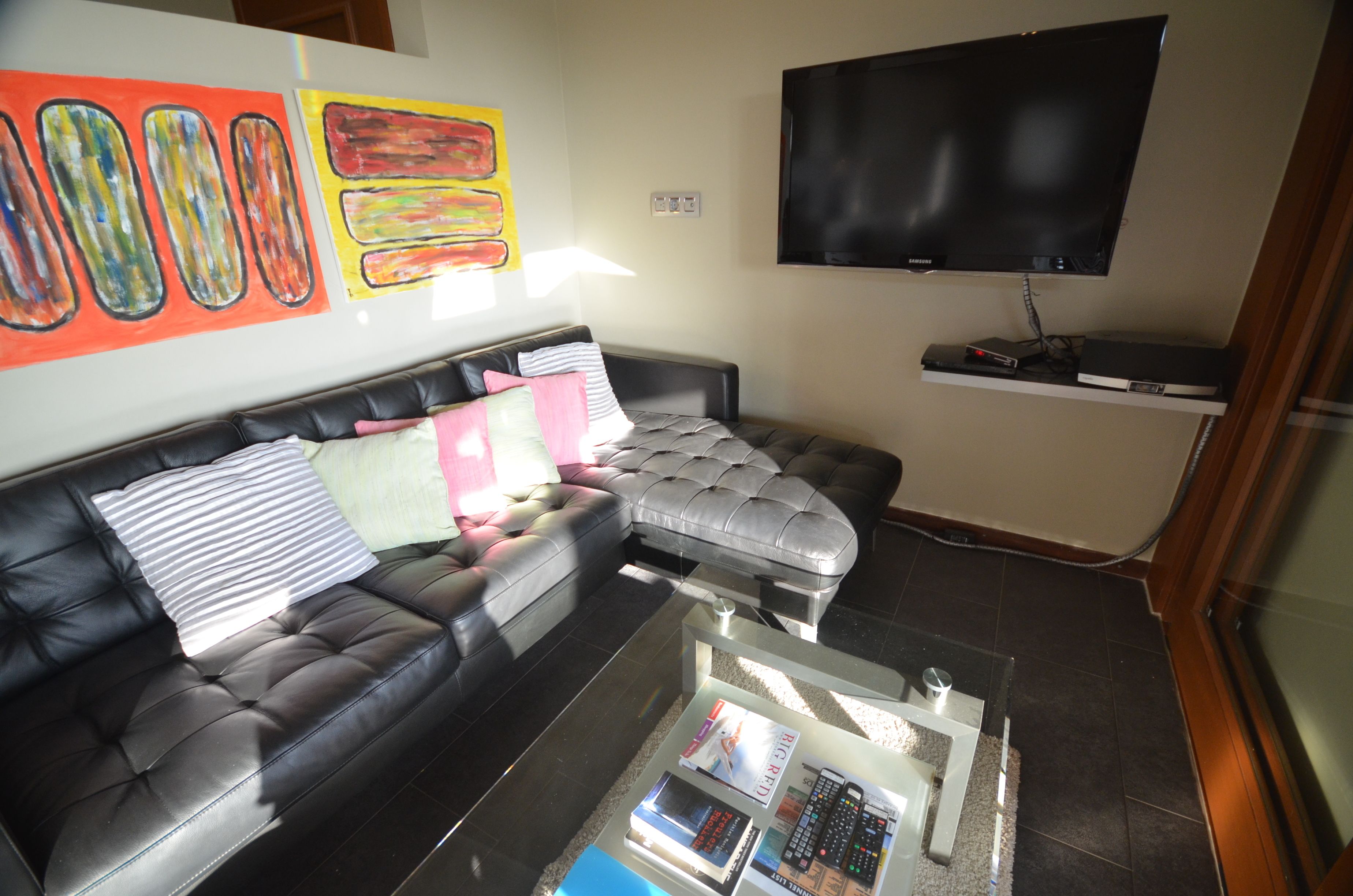 Una sala de estar con un sofá de cuero negro, una mesa de centro de vidrio con revistas, un televisor de pantalla plana montado en la pared y arte abstracto en la pared.