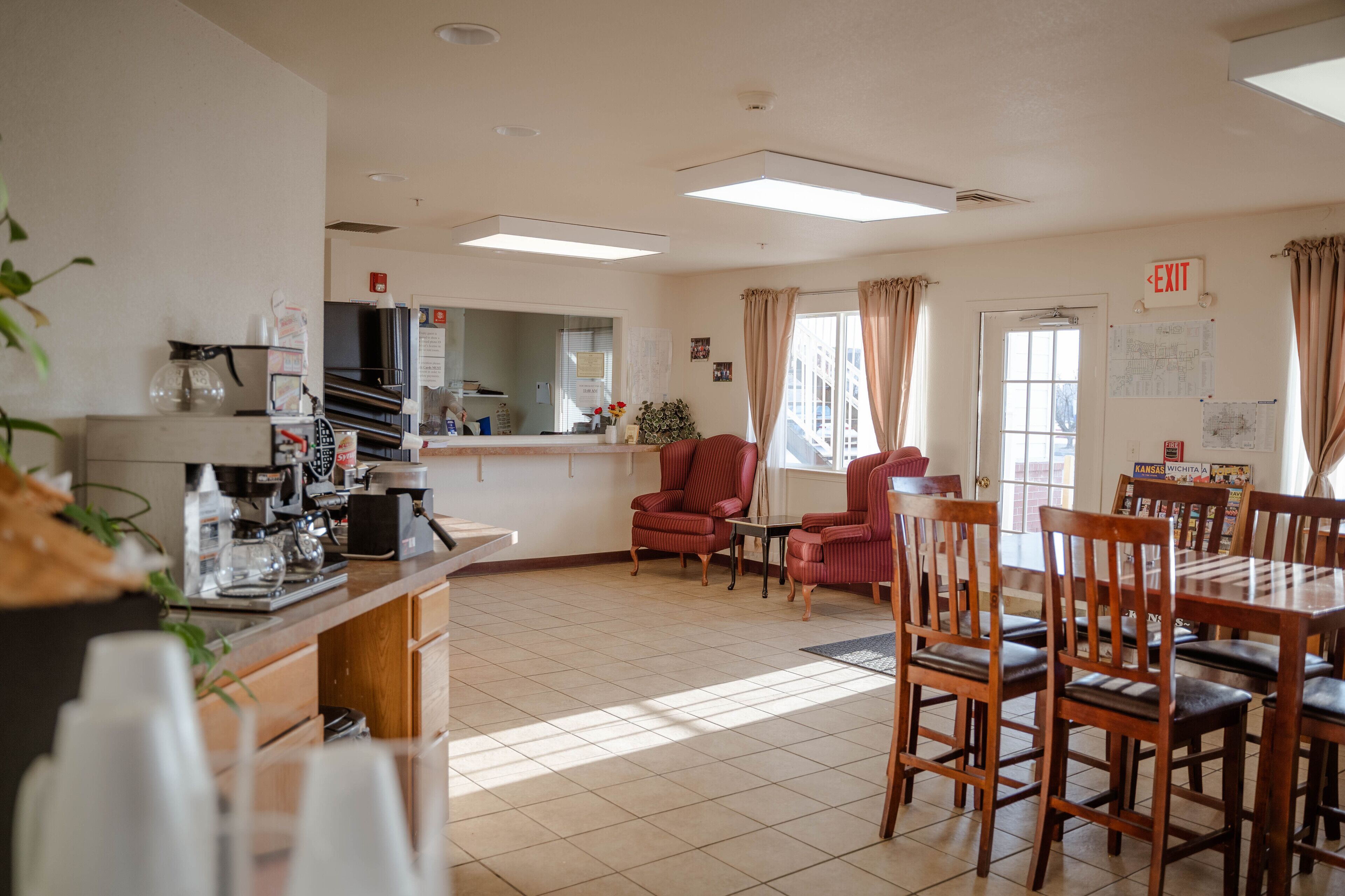 Un grand espace de salle à manger avec une longue table et plusieurs chaises, un coin cuisine équipé d’une machine à café, et une grande fenêtre avec des rideaux.