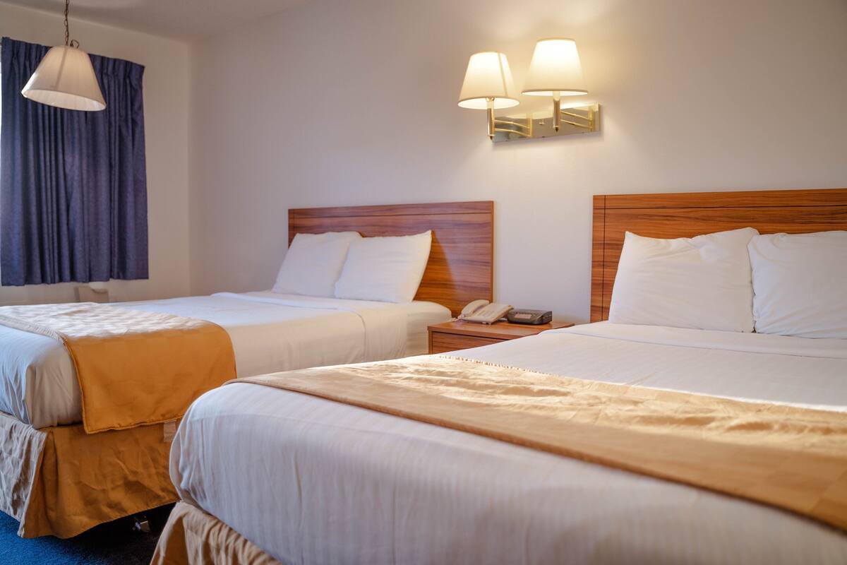 Habitación de hotel con dos camas, cabecero de madera, mesita de noche con lámpara y cortina.