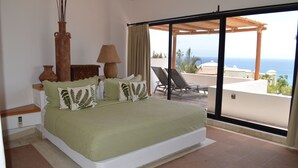 Un dormitorio con una cama grande, una mesita de noche, una lámpara y vistas al océano a través de un ventanal corredera.
