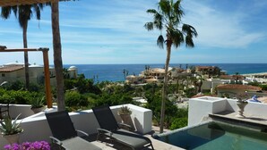 Un área junto a la piscina con sillones de descanso, una piscina pequeña y vista al océano y edificios en el fondo.