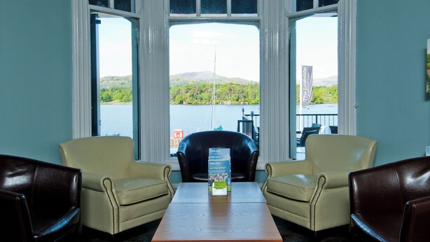 A room with a large bay window overlooking a lake, two leather chairs, a wooden table, and a banner advertising a lakeside restaurant. - YHA Ambleside - Hostel (Ambleside)