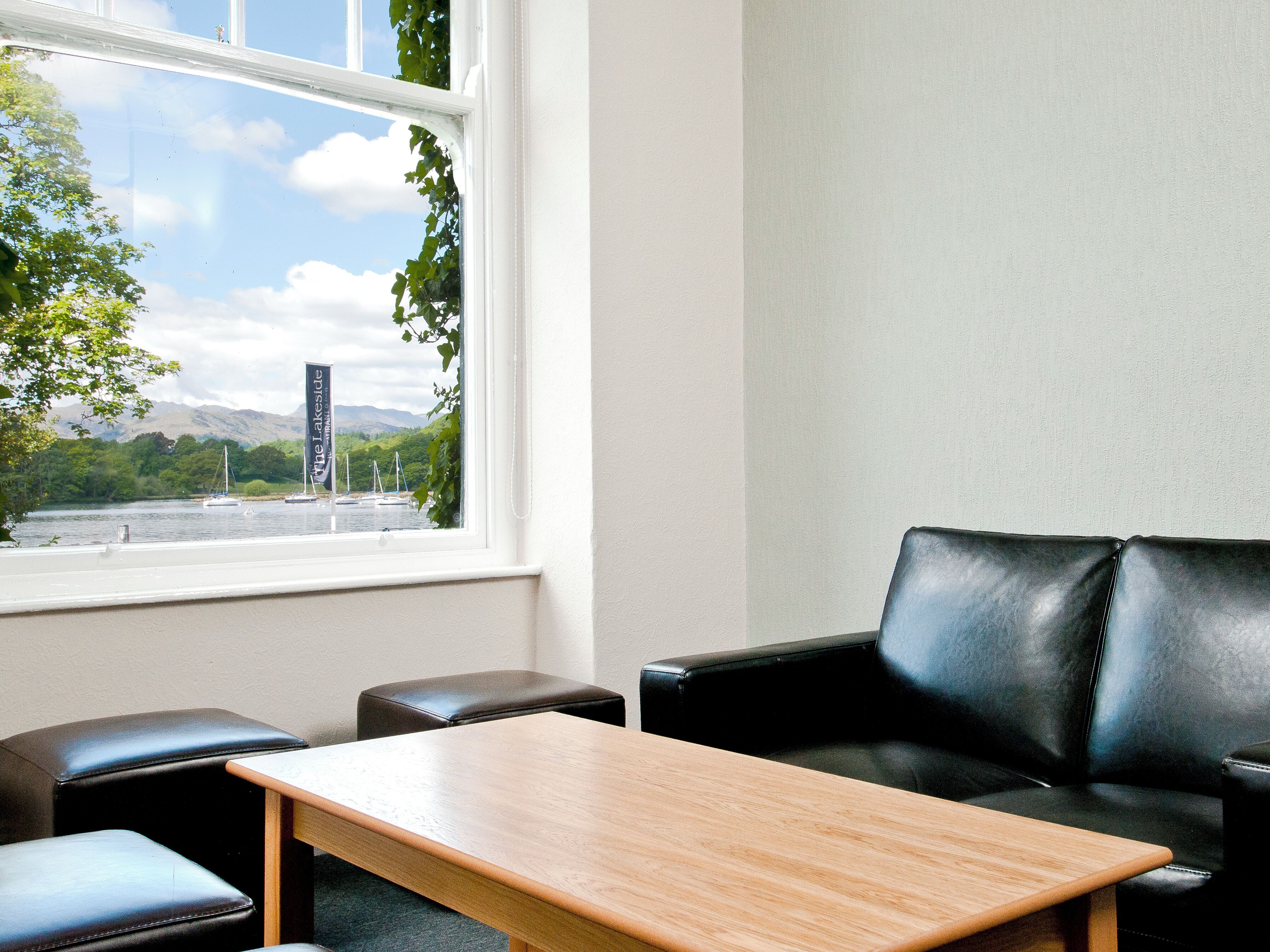 Una habitación con una mesa de madera, dos sillas de cuero negro y una ventana que ofrece vistas a un lago con barcos y vegetación.
