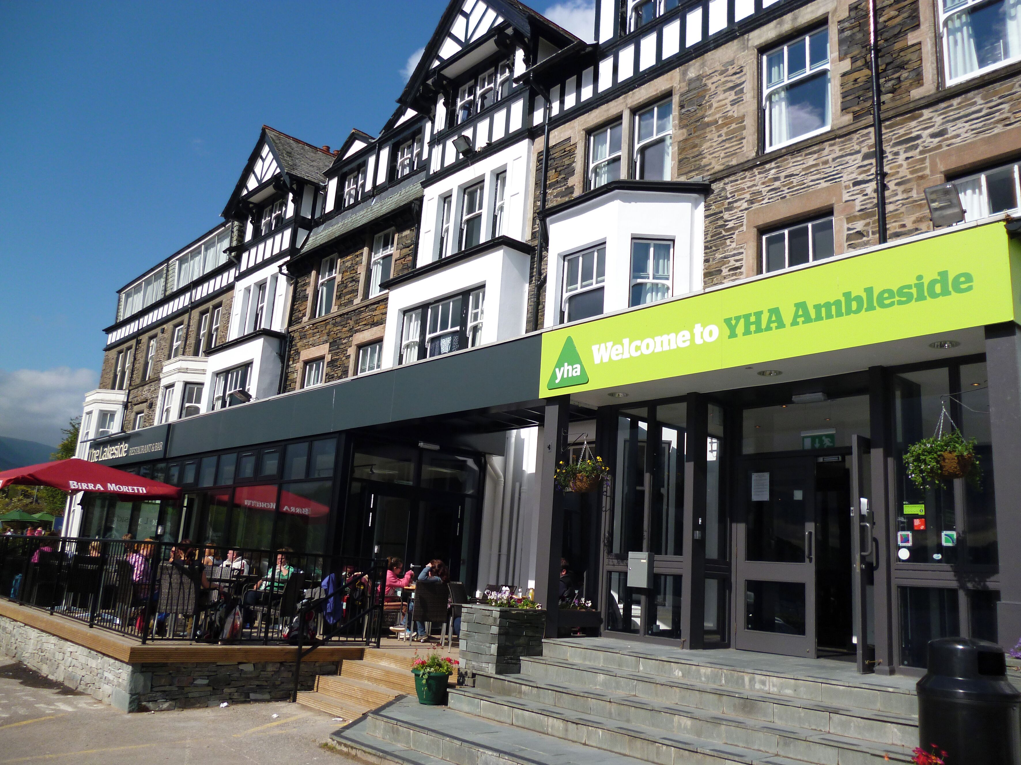 A building with a sign reading "Welcome to YHA Ambleside" and a restaurant named "Birra Moretti" with outdoor seating.