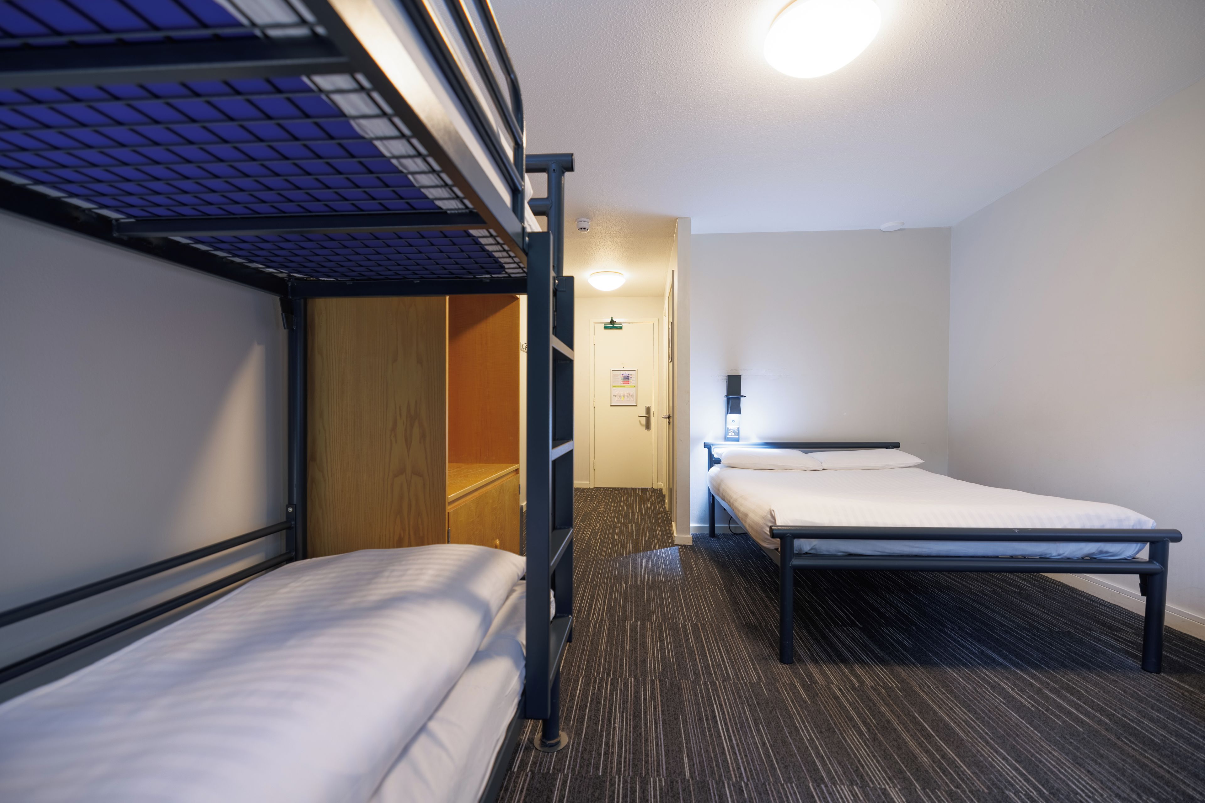 A hotel room with bunk beds, a wooden wardrobe, a door, a wall-mounted light, and a carpeted floor.
