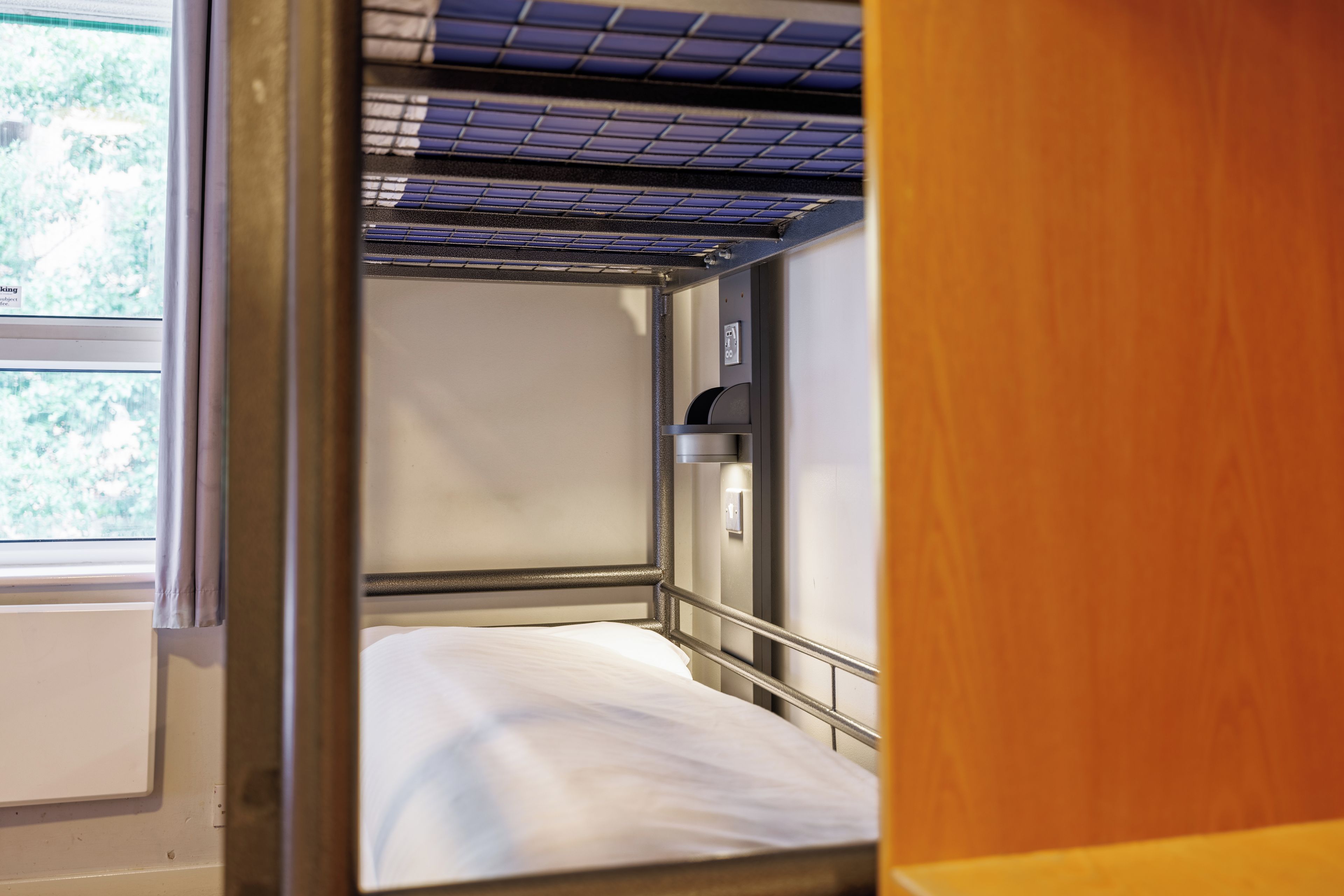 Habitación pequeña con cama individual, ventana, puerta y un estante con lámpara.
