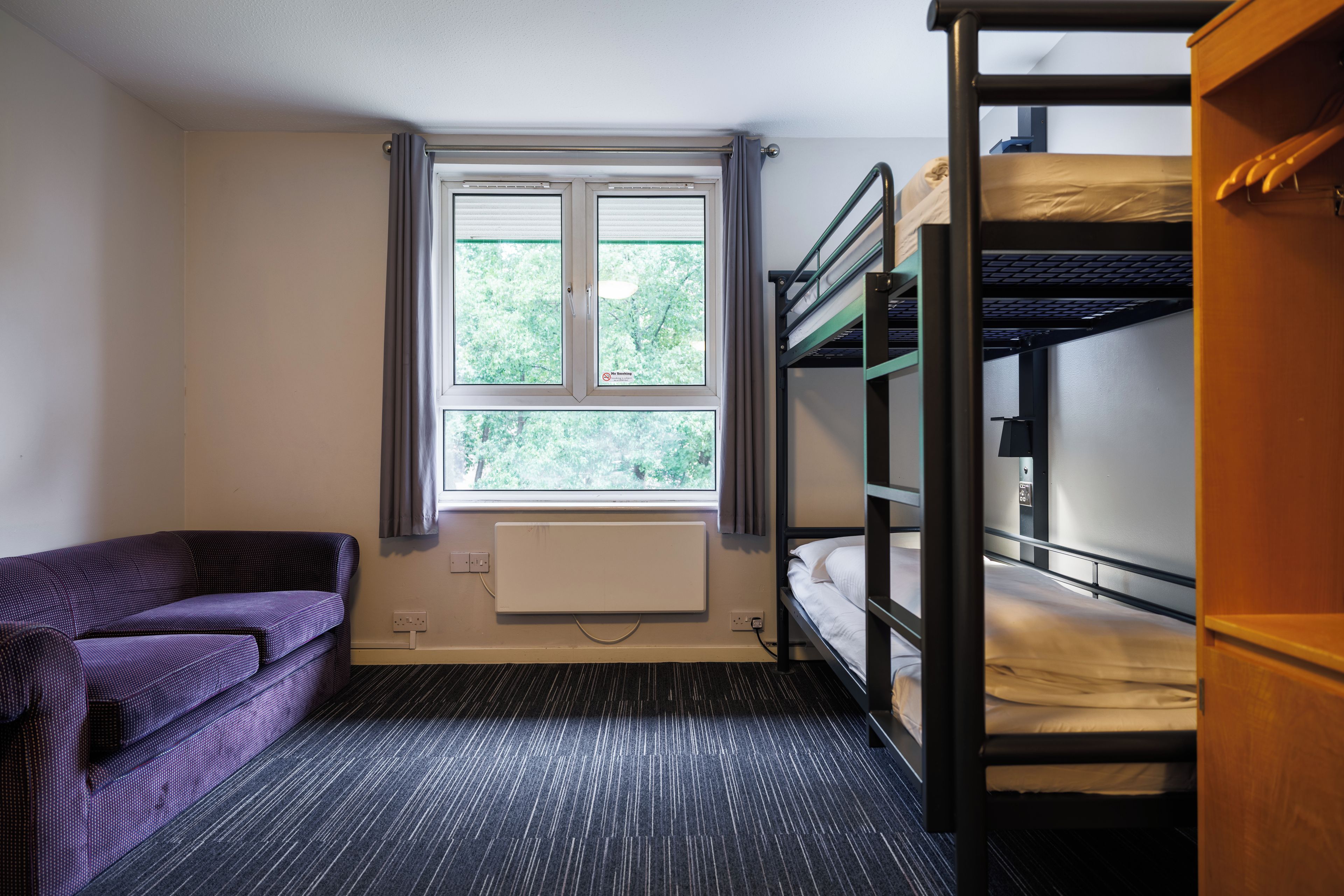A dormitory room with bunk beds, a window with curtains, a radiator, and a purple sofa.