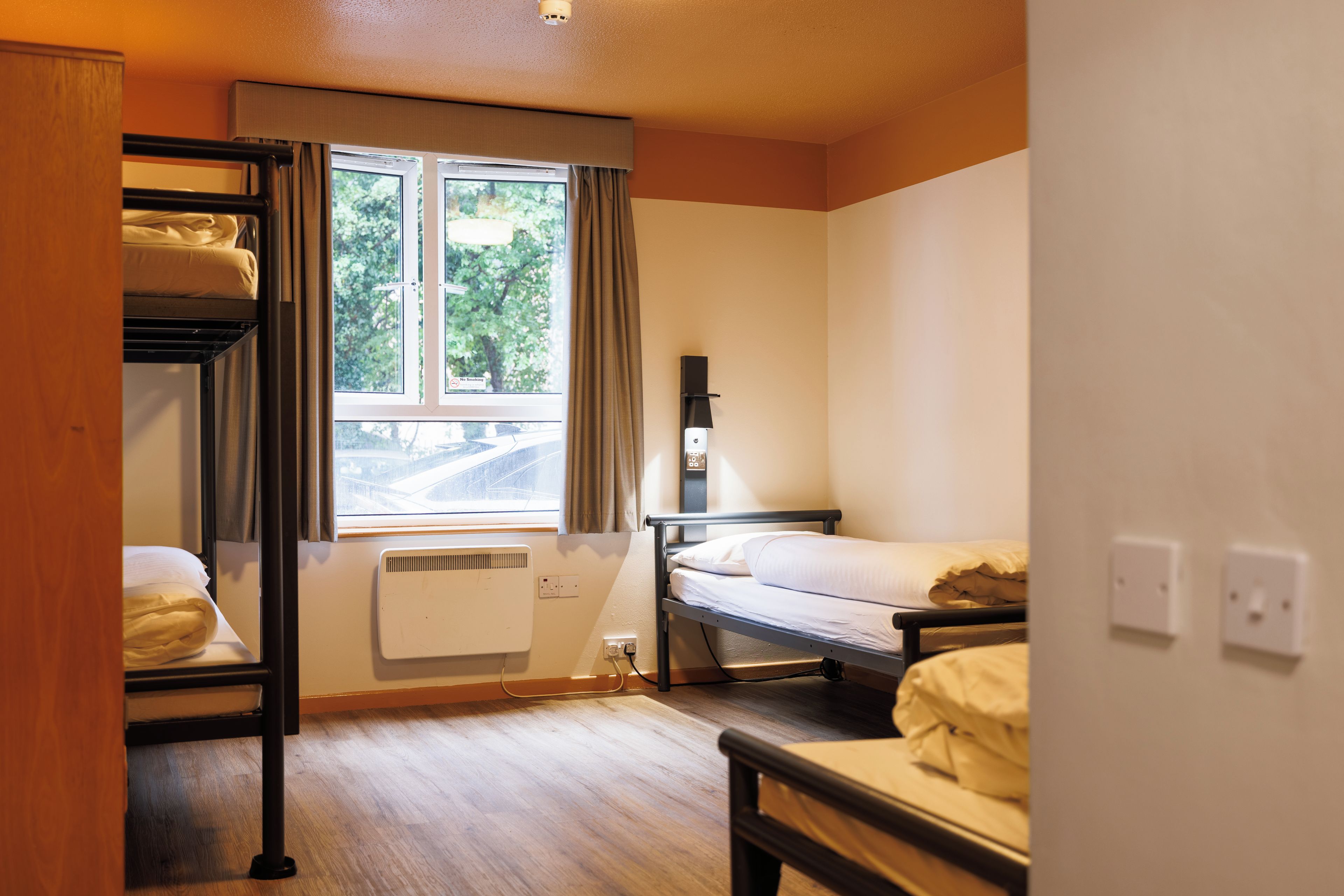 Une chambre de dortoir avec des lits superposés, une fenêtre avec des rideaux, un radiateur et un luminaire fixé au mur.