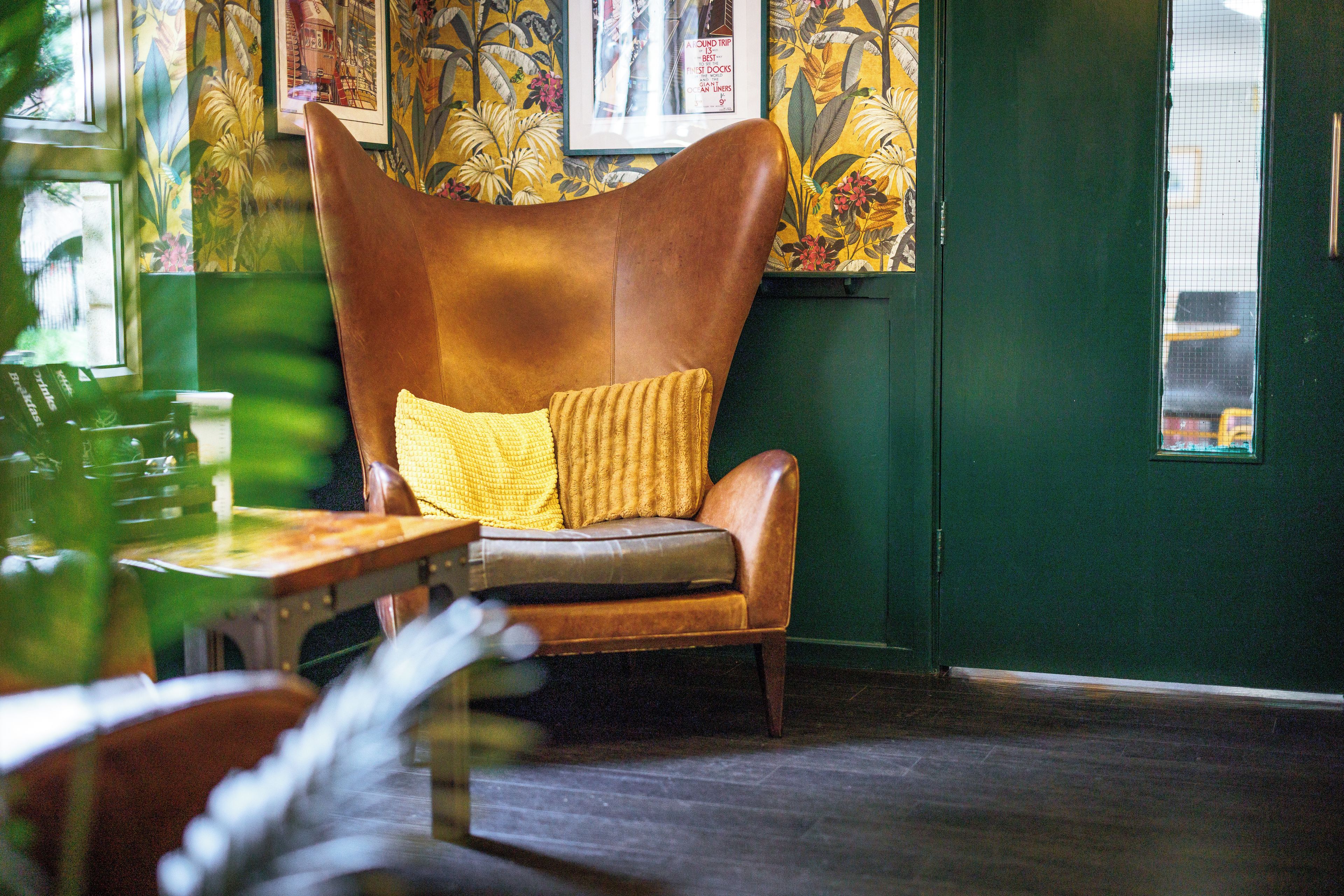 A room with a brown leather armchair with a yellow cushion, a wooden table, and a green wall with floral wallpaper.